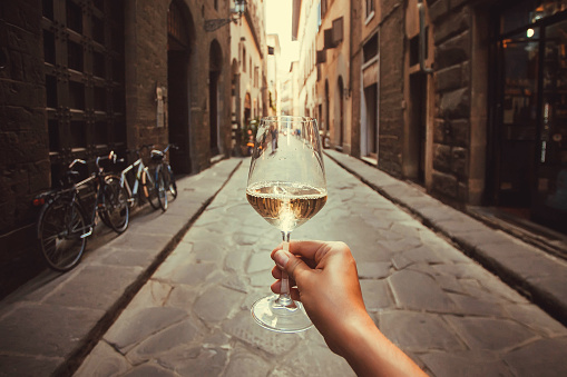 Wine glass in hand of happy tourist walking past historical houses of Florence, Tuscany. Ancient streets in Italy.