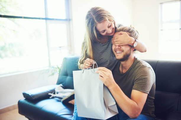 Happy birthday, handsome Shot of a young woman surprising her husband with a gift at home boyfriend stock pictures, royalty-free photos & images