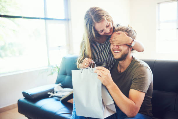 Happy birthday, handsome Shot of a young woman surprising her husband with a gift at home gift for girlfriend stock pictures, royalty-free photos & images
