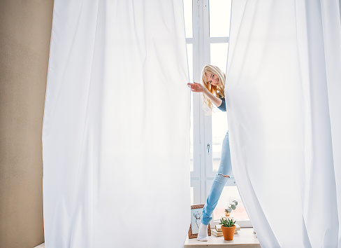 Beautiful blonde girl in jeans standing behind white curtains. The blonde woman is looking out of a curtain