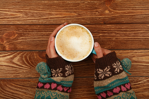 Close up two woman hands hold and hug big full cup of latte cappuccino coffee with milk froth over brown wooden table, elevated top view, directly above