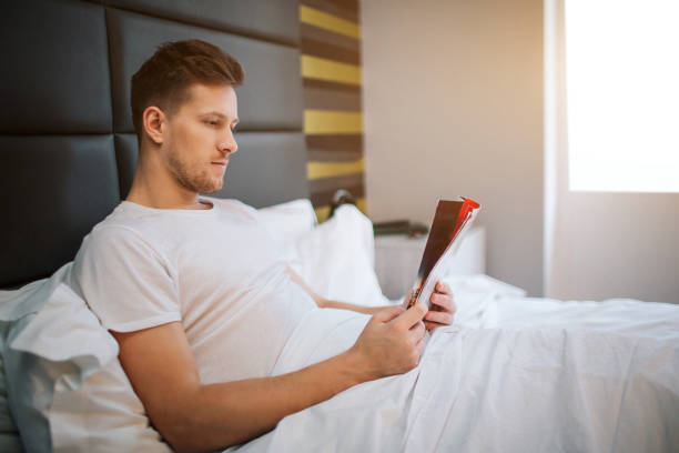 istock Young handsome man read in bedroom. Concentrated. Daylight. He covered with white blanket. 1135127209