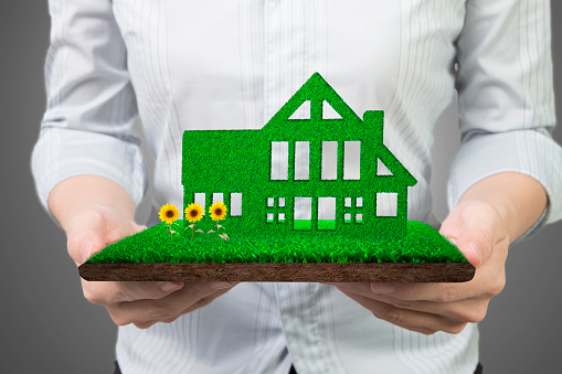 Green energy supply and solution concept, woman hands holding green house on grass land with mud and sunflowers, front view, isolated on gray background.