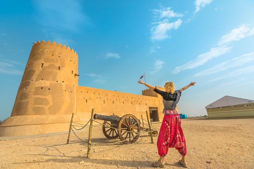 Travel in North of Qatar. Carefree woman at Al Zubara Fort, a historic Qatari military fortress, Middle East, Arabian Peninsula. Blonde tourist traveler in front of cannon in old castle, Persian Gulf.