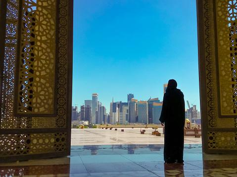 Woman with abaya dress looks at views of modern skyscrapers of Doha West Bay skyline outdoors State Grand Mosque in Doha, Qatar, Middle East, Arabian Peninsula. Sunny day with blue sky.