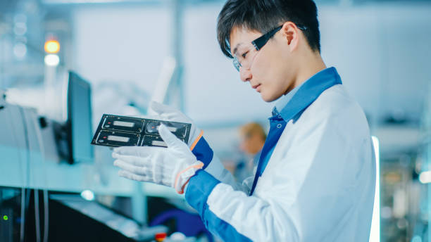 istock High-Tech Factory: Quality Control Engineer Checks Electronic Printed Circuit Board it for Damages. In the Background Assembly Line for PCB with Surface Mount Pick and Place Technology. 1135140556