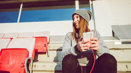 Young woman sitting on a stadium seat, listening to music over headphones