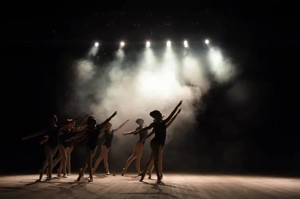 Photo of Ballet class on the stage of the theater with light and smoke. Children are engaged in classical exercise on stage.