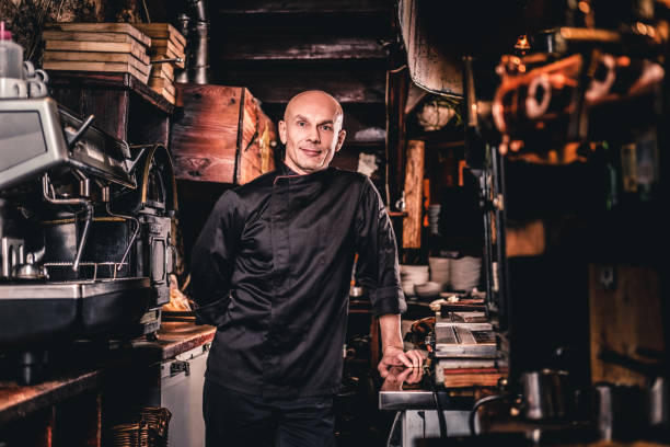 istock Confident chef wearing uniform leaning on a table and looking at a camera in a restaurant kitchen. 1135148475
