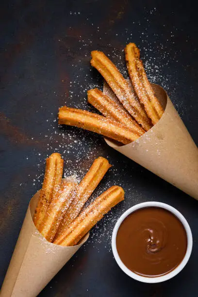 Traditional churros sticks in paper bag with sugar powder cinnamon and bowl of chocolate dip on dark table top view