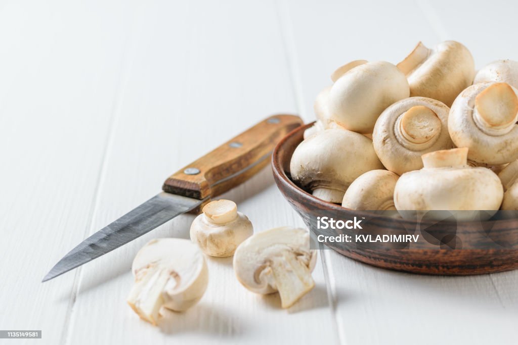 A clay bowl with mushrooms, a knife and pieces of mushrooms on a white wooden table. Vegetarian cuisine. The view from the top. Flat lay. A clay bowl with mushrooms, a knife and pieces of mushrooms on a white wooden table. Vegetarian cuisine. Natural plant food. The view from the top. Agaricus Stock Photo