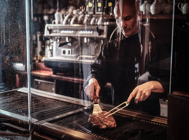 chef che cucina deliziosa bistecca di manzo in cucina, in piedi dietro un bicchiere protettivo in un ristorante. - silverware food and drink industry food service occupation dinner foto e immagini stock