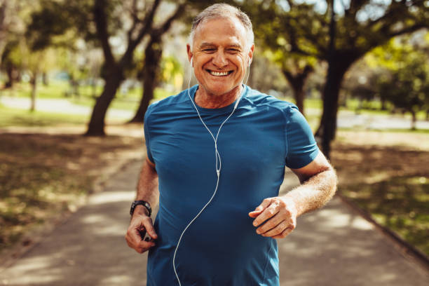 Senior man working out for good health Portrait of a senior man in fitness wear running in a park. Close up of a smiling man running while listening to music using earphones. fitness stock pictures, royalty-free photos & images