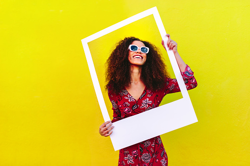Pretty woman in a red dress and sunglasses standing against a yellow wall and holding a large empty frame. Beautiful female model posing with empty picture frame.