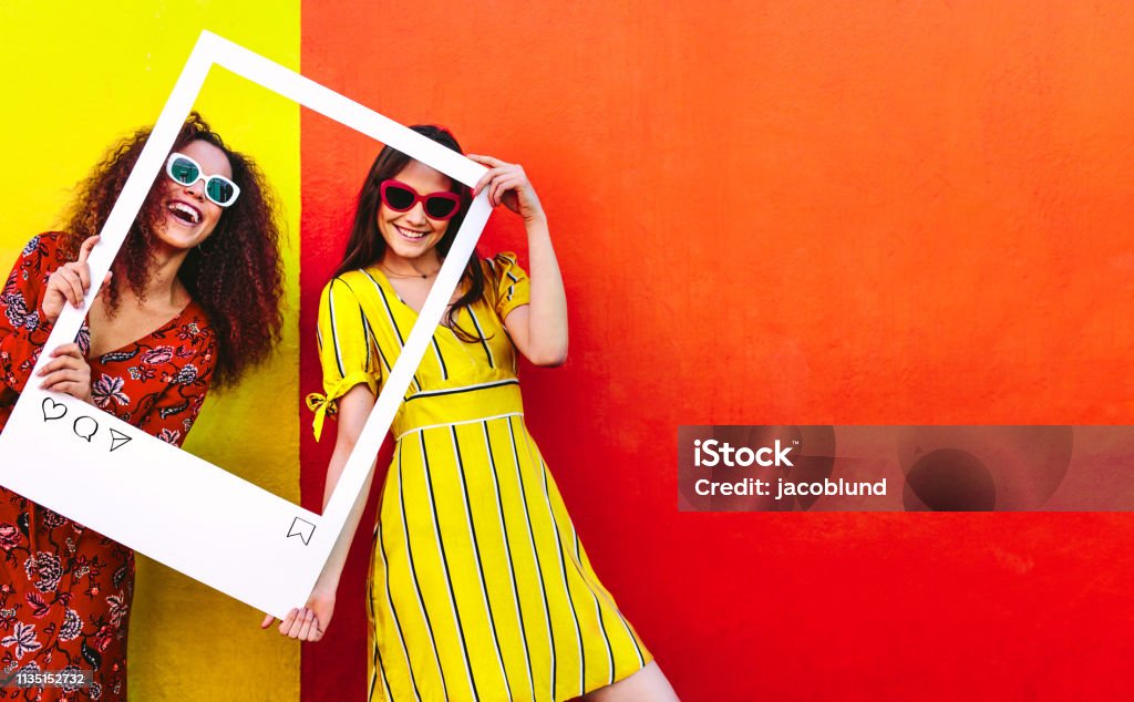 Friends posing for their social media post photo Portrait of two women holding a blank photo frame in hand and smiling. Girls wearing sunglasses standing against red and yellow colored wall. Social Media Stock Photo