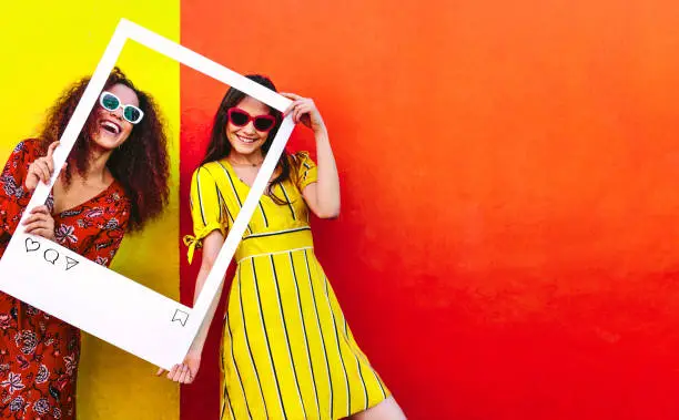 Portrait of two women holding a blank photo frame in hand and smiling. Girls wearing sunglasses standing against red and yellow colored wall.