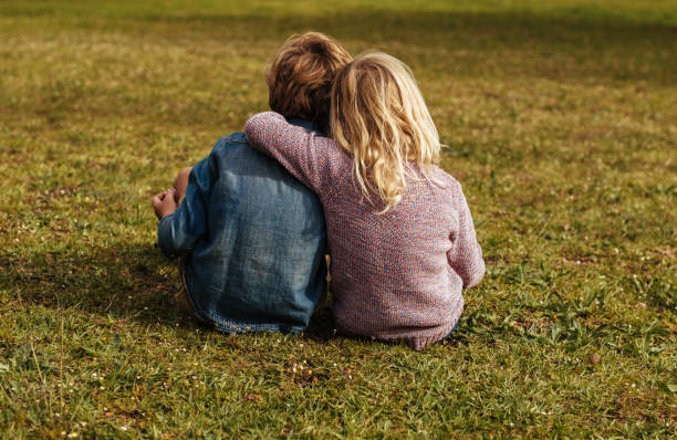 Siblings sitting on the grassy lawn Rear shot of siblings sitting on the grass. Little girl sitting with her brother putting her arm on his shoulder at park. sister stock pictures, royalty-free photos & images