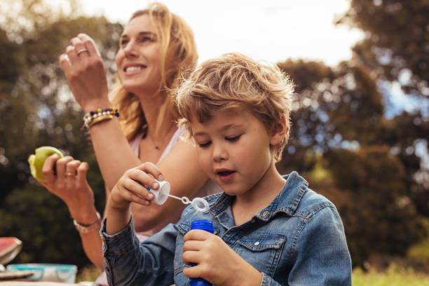 Little boy makes soap bubbles with her mom sitting by in the park. Small kid on picnic with his mother at the park.