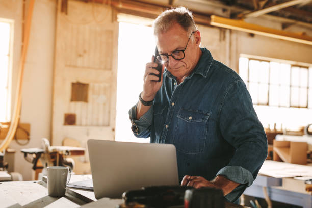 Senior male carpenter working in his workshop Senior man talking on cell phone and using laptop on work table. Mature carpenter working on laptop and answering phone call in his carpentry workshop. working class stock pictures, royalty-free photos & images