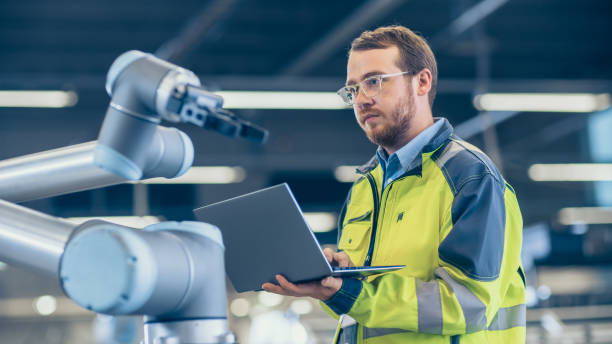 in fabbrica: l'ingegnere dell'automazione utilizza laptop per la programmazione di un braccio robotico. nuova era nell'industria manifatturiera automatica. - opperated foto e immagini stock