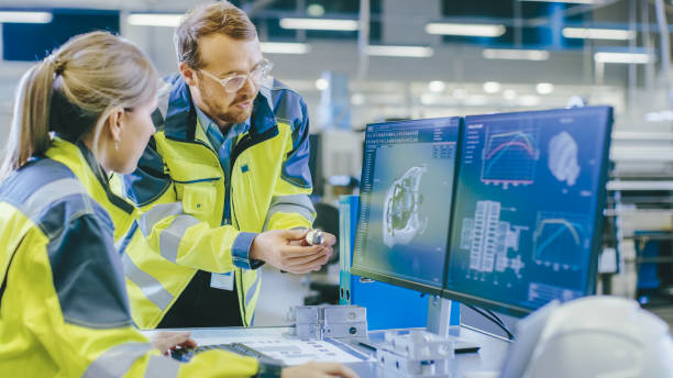 istock At the Factory: Male Mechanical Engineer Holds Component and Female Chief Engineer Work on Personal Computer, They Discuss Details of the 3D Engine Model Design for Robotic Arm. 1135159598