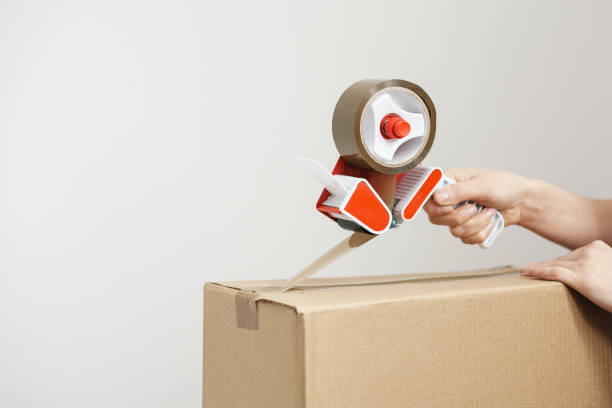 young beautiful brunette girl in a white t-shirt packs cardboard boxes with a dispenser and adhesive scotch adhesive tape. the concept of moving to a new home. - packaging packing adhesive tape box imagens e fotografias de stock