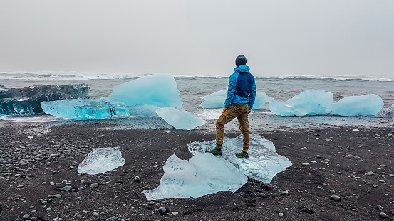 A young man wearing a blue jacket stands on a smaller ice berg, washed to the beach by the sea. Black sand beach. Blue shades of ice. Sea drifting the ice bergs. Global warming effects.