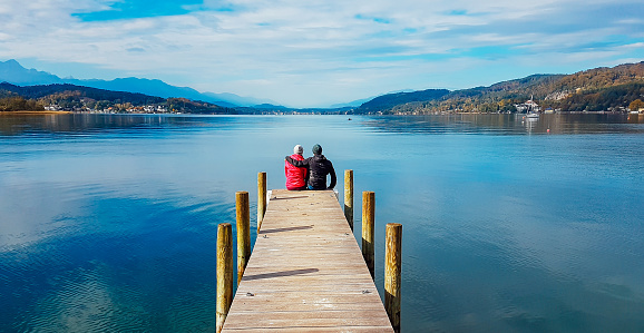 Couple sitting and hugging at the pier by Wörtersee, Pörtschach, Austria. Beautiful lake landscape, surrounded by Alps. This lake is natural drinking water tank. Love is in the air. Romantic moments.