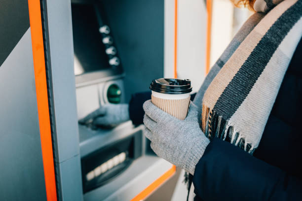 Close-up of young woman in warm clothing Close-up of young woman in warm clothing holding coffee and using an ATM on city street in winter day. exchange teller stock pictures, royalty-free photos & images