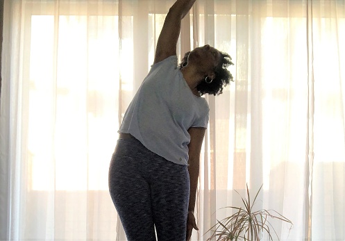 A mature woman of African descent doing a standing side bend yoga pose in her home during the day, sunlight in the background creating a silhouette