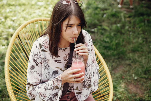 Stylish hipster boho girl drinking strawberry smoothie in glass jar with metal reusable straw at street food festival. Happy woman in sunglasses with healthy drink. Zero waste