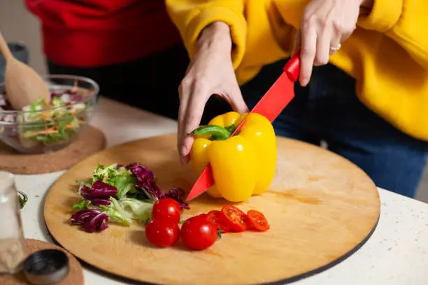 Woman in yellow sweater holding a sharp red knife while cutting big yellow paprika. Cherry tomatoes and lettuce on the cutting board near