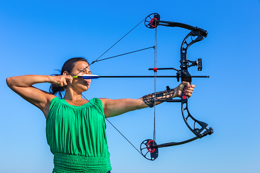 Young black haired colombian woman aiming arrow of compound bow in blue sky. On this sunny day in summer season there was a beautiful blue sky. The female person was training and learning to shoot with bow and arrow.