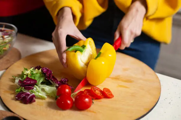 Close up of hands of woman using red sharp knife while cutting yellow fresh paprika on the cutting board