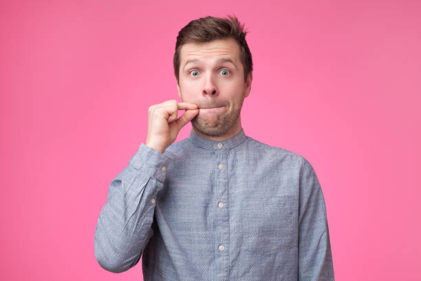 Young man showing a sign of closing mouth and silence gestur Young man showing a sign of closing mouth and silence gesture doing like closing his mouth with a zipper on isolated pink background silence stock pictures, royalty-free photos & images
