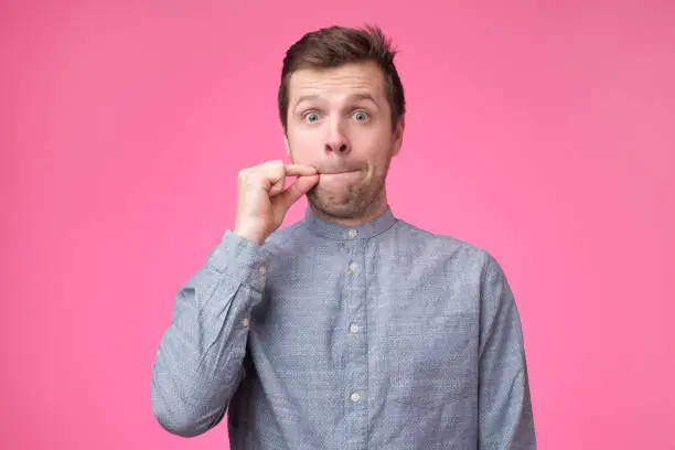 Young man showing a sign of closing mouth and silence gesture doing like closing his mouth with a zipper on isolated pink background