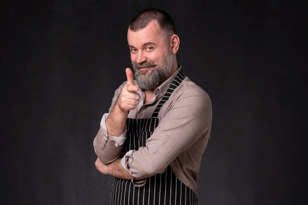 istock Mature bearded man in apron pointing with finger at camera 1135215362
