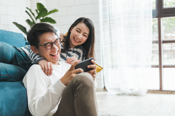 家庭で一緒に携帯電話でゲームをプレイするアジアのカップルの男性と女性、アジアのカップルの家族のライフスタイルの概念 - portable player ストックフォトと画像