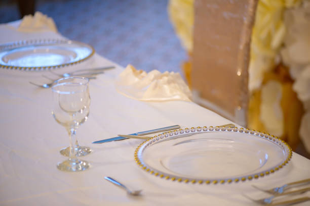 Table sitting arrangement at a formal event or fine dining restaurant featuring transparent plates with golden details, glassware and silverware in the order of use set against a paper flowers wall.