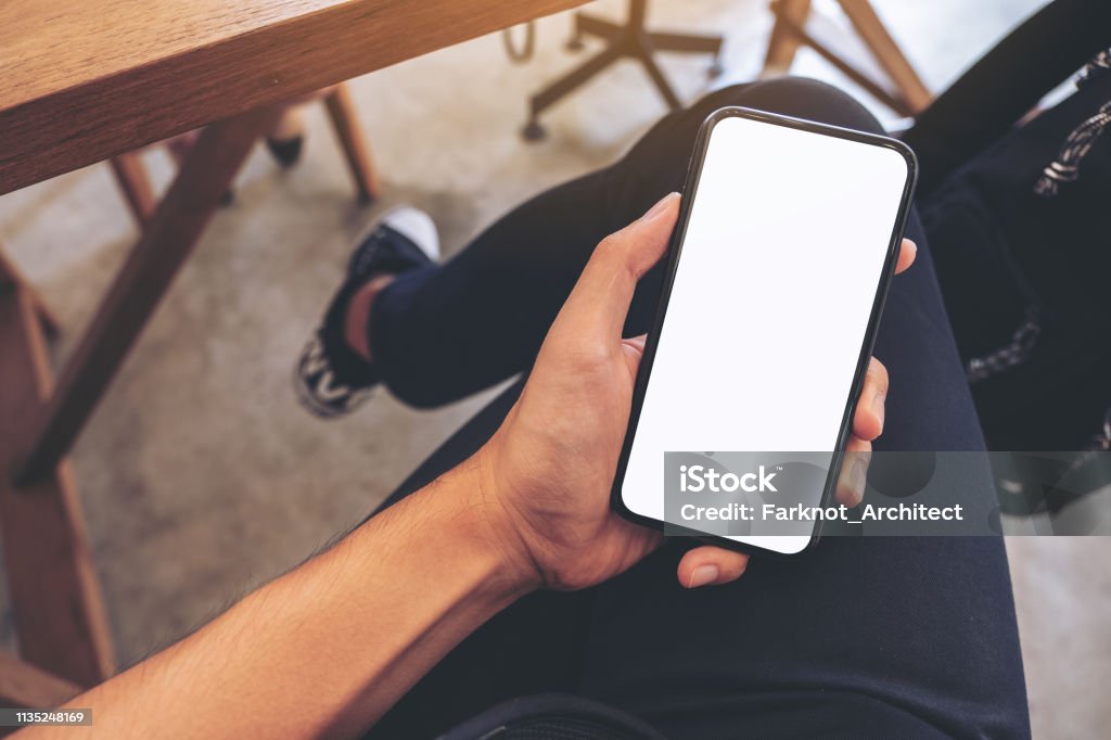 Top-Ansicht Mockup-Bild von der Hand eines Mannes hält weißes Handy mit blankem Desktop-Bildschirm auf dem Oberschenkel, während im Café sitzen - Lizenzfrei Telefon Stock-Foto