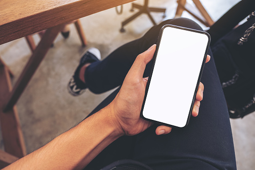Top view mockup image of a man's hand holding white mobile phone with blank desktop screen on thigh while sitting in cafe