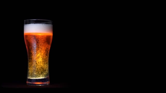 Glass of golden craft beer with white foam and moving bubbles isolated at black background