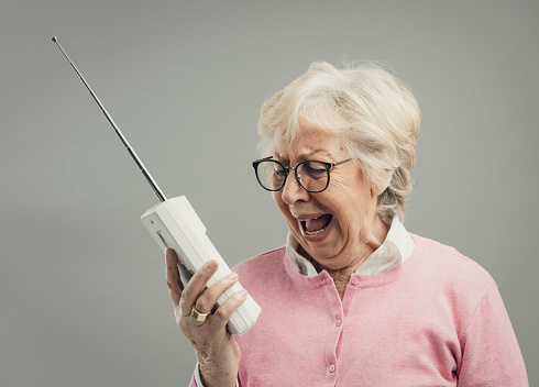 Frustrated senior woman using an old cordless telephone, she is shouting out loud and talking to a deaf person