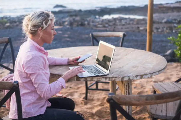 digital nomad concept with blonde free beautiful caucasian middle age woman working at the laptop using cellular phone too sitting on a wood table near the ocean - freelance free to work everywhere with technology