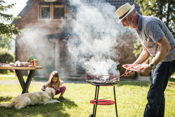 hombre maduro preparando barbacoa para su nieto en el patio trasero. el enfoque está en el hombre. - sausage barbecue grill barbecue cooking fotografías e imágenes de stock