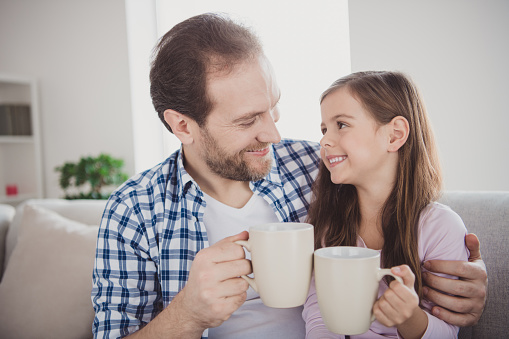 Close-up portrait of his he her she nice cute lovely attractive cheerful peaceful pre-teen girl handsome bearded dad daddy sitting on divan drinking cacao in modern light white interior room indoors.