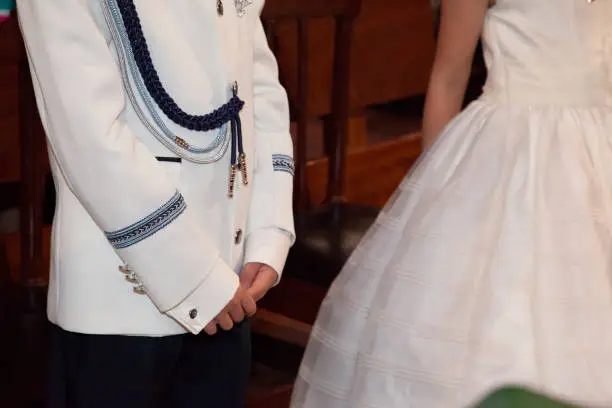 detail of the intertwined hands of a child who is receiving the first communion. You can see her nice white jacket suit with blue decorations, in the background you can see the costume of a girl who is also taking communion and who is at her side.