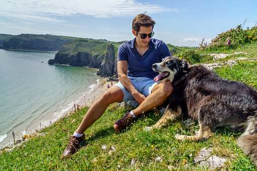 Single Hispanic man lifestyle portrait at beach sitting on headland at Skrinkle Haven on the Pembrokeshire Coast with his dog