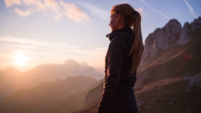 Woman standing on top of mountain, enjoying breathtaking view at sunset