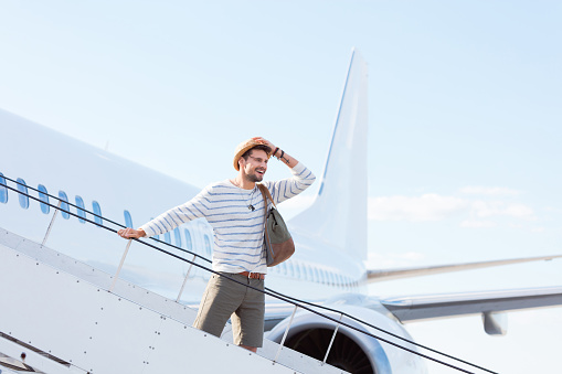 Handsome young man holding his hat and standing on the stairway of an airplane. Man disembarking a flight.